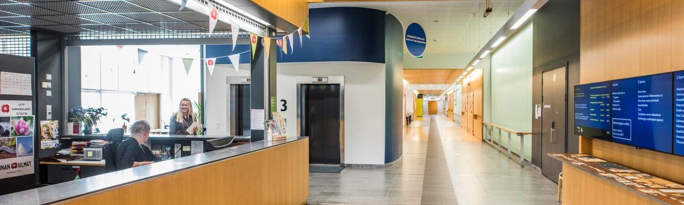 Edessä vasemmalla vastaanottotiski, jonka yläpuolelle pieniä viirejä. Tiskin takana kaksi hissiä ja kerrosnumero 3. Hissien oikealta puolelta jatkuu pitkä ja valoisa käytävä, jonka varrella ovia.